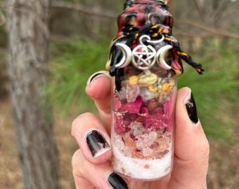 May include: A small glass bottle with a black, red, and yellow patterned exterior. The bottle is filled with pink and white crystals, dried flowers, and herbs. A silver charm with a triple moon and pentagram hangs from the bottle's neck.