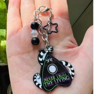 May include: A black and white striped charm with a green, purple, and white Ouija board design. The charm has the text "Never Trust The Living" on it. The charm is attached to a silver chain with black and clear beads and two silver star charms.