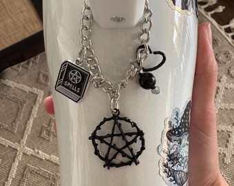 May include: A white ceramic mug with a silver chain charm. The charm has a black pentagram, a black book with the word "Spells" on it, and a black bead. The mug has a holographic finish and a sticker of a toadstool.