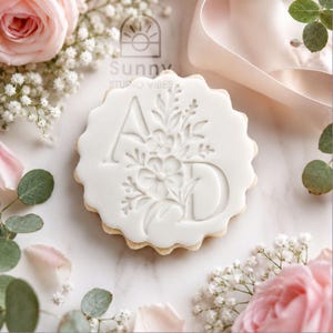 May include: A round white biscuit with scalloped edges featuring an embossed design of the letter A and the letter D intertwined with a detailed floral arrangement of leaves and small blossoms centred on the smooth surface of the icing.