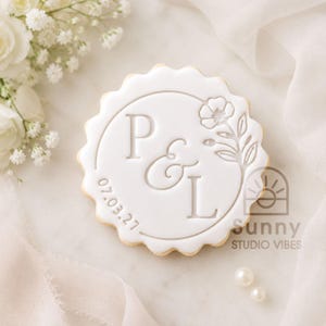 May include: A round, scalloped-edge white biscuit featuring embossed lettering P & L, a floral sprig design, and the date 07.03.27. The biscuit is decorated with smooth white icing and displays a clean, centred layout with a circular border.