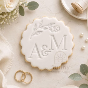 May include: A round, scalloped-edge biscuit with white icing featuring the embossed letters A & M and a floral sprig design. The biscuit is positioned near two gold rings, white flowers, and a spool of ribbon, with the text Sunny Studio Vibes.