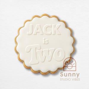 Può includere: Un biscotto rotondo con glassa bianca e bordo smerlato. Il testo "JACK is Two" è impresso sulla glassa. Il biscotto ha un bordo marrone dorato. Il logo Sunny Studio Vibes si trova in basso a destra.