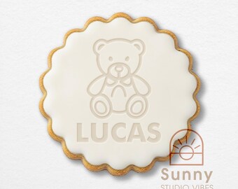 May include: A round, scalloped-edge cookie with a smooth white icing finish. The center features an embossed graphic of a seated teddy bear above the name LUCAS in capital letters. A small logo with the text Sunny STUDIO VIBES is visible.
