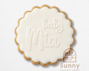 May include: A round scalloped cookie with white icing featuring the embossed text baby Mia with a small heart above the letter i. The cookie has a golden brown edge and a smooth surface texture. The logo Sunny Studio Vibes is in the corner.