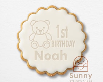 May include: A round scalloped cookie with white icing featuring an embossed teddy bear icon, the text 1st BIRTHDAY, and the name Noah. The cookie has a golden-brown baked edge and includes a small logo in the corner reading Sunny STUDIO VIBES.