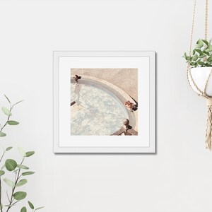 May include: A framed photograph of a round pool with a light blue surface. The pool is surrounded by a light brown stone edge. The pool is partially filled with water and there are two shadows in the water.