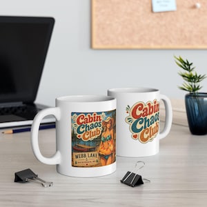 May include: Two white ceramic mugs featuring a colorful graphic design. One mug displays the text Cabin Chaos Club, Webb Lake, Hackensack, MN, with an illustration of a person in a swimsuit and a lake scene. The other mug shows the text Cabin Chaos Club.