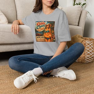May include: A heather gray t-shirt featuring a graphic print of a person in a bikini and sun hat holding an inflatable ring. The text reads Cabin Chaos Club, Webb Lake, and Hackensack, MN, with illustrations of a boat and a wooden cabin structure.