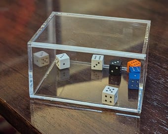 May include: A clear acrylic box with six dice inside. The dice are white, black, blue, and orange. The box is on a wooden surface.