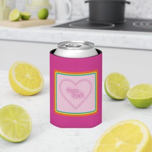 Puede incluir: Un enfriador de latas rosa con un diseño de corazón y el texto "Casa Bach". El enfriador tiene un borde de arcoíris y sostiene una lata plateada. Limones y limas en rodajas están esparcidos alrededor del enfriador.
