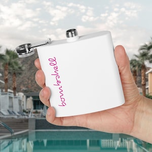 Puede incluir: Un frasco blanco con tapa y bisagra plateadas, sostenido en una mano. La palabra "bombshell" está escrita en rosa en la parte delantera. El fondo muestra una piscina y palmeras.