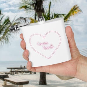 May include: A white flask with a silver cap and hinge. The flask has a pink heart design with the words "Casa Bach" inside. The background shows a beach scene with palm trees and the ocean.
