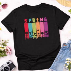 May include: Black t-shirt with the word "SPRING" in rainbow colors. Below the word are vertical stripes of color with silhouettes of flowers, birds, and plants. The shirt is made of a soft material.