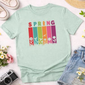 May include: A light green t-shirt with the word "SPRING" in colorful letters. Below the word are vertical stripes of red, blue, green, orange, pink, and purple, each with a floral or bird graphic. The shirt is paired with denim shorts and sneakers.