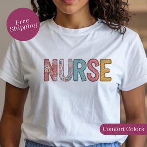 Puede incluir: Camiseta blanca con la palabra "NURSE" en letras coloridas estilo patchwork. Las letras son rosas, azules y amarillas, con estampados florales y a rayas. La camiseta tiene mangas cortas.