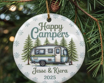 Op de afbeelding: Ronde witte keramische kerstversiering met de tekst "Happy Campers" en een illustratie van een vintage camper. Het bevat de namen "Jesse & Kiera" en het jaar "2025". Versierd met sneeuwvlokken en dennenbomen.