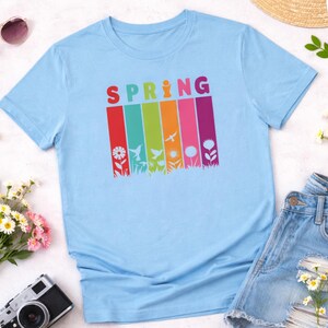 May include: Light blue t-shirt with the word "SPRING" in colorful letters. Below the word are vertical stripes in red, green, yellow, orange, pink, and purple, each with a spring-themed graphic, including flowers and birds.