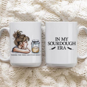 Può includere: Due tazze in ceramica bianca con testo e illustrazioni neri. Una tazza presenta una donna con un barattolo di pasta madre e la scritta "FEEDING THIS THING LIKE IT PAYS RENT". L'altra tazza dice "IN MY SOURDOUGH ERA" con illustrazioni di grano.