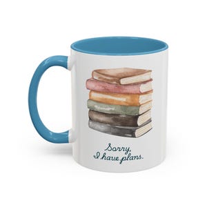 Puede incluir: Taza de cerámica blanca con asa e interior azules. La taza presenta una ilustración de acuarela de una pila de libros en tonos marrón, rosa, naranja, verde y negro. El texto "Sorry, I have plans." está impreso en la taza.
