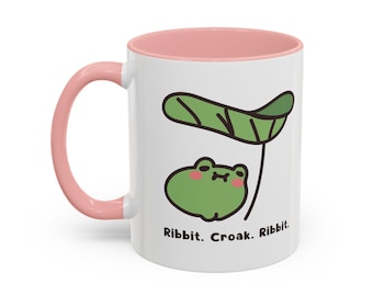May include: White ceramic mug with a pink handle and interior. The mug features a cartoon frog under a leaf, with the text "Ribbit. Croak. Ribbit." in black. The frog is green with pink cheeks.