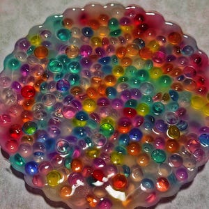 May include: A colorful, round resin coaster filled with vibrant, translucent water beads. The beads are in a variety of colors, including red, orange, yellow, green, blue, and purple. The coaster has a wavy edge.