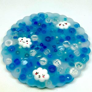 May include: A round, blue resin coaster with a scalloped edge. The coaster is filled with blue beads and three white cloud-shaped charms with smiling faces. The design evokes a whimsical, weather-themed aesthetic.