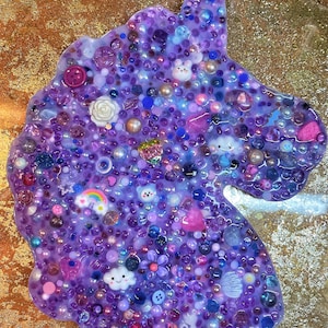 May include: A purple unicorn-shaped resin art piece, encrusted with a variety of embellishments. The design includes buttons, beads, pearls, and small decorative elements like clouds, rainbows, and flowers. The background has a gold glitter effect.