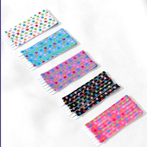 May include: Five colorful bobby pins with polka dot patterns. The pins feature a variety of colors, including white, blue, purple, black, and pink, each adorned with a different color of polka dots. The bobby pins are arranged diagonally on a white surface.