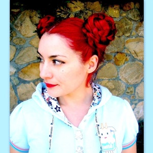 Puede incluir: Una persona con cabello rojo brillante peinado en dos mo&ntilde;os trenzados. Lleva una sudadera con capucha azul claro con un gr&aacute;fico de dibujos animados y ribetes estampados. El fondo es una pared de piedra.