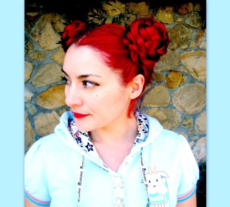Puede incluir: Una persona con cabello rojo brillante peinado en dos mo&ntilde;os trenzados. Lleva una sudadera con capucha azul claro con un gr&aacute;fico de dibujos animados y ribetes estampados. El fondo es una pared de piedra.