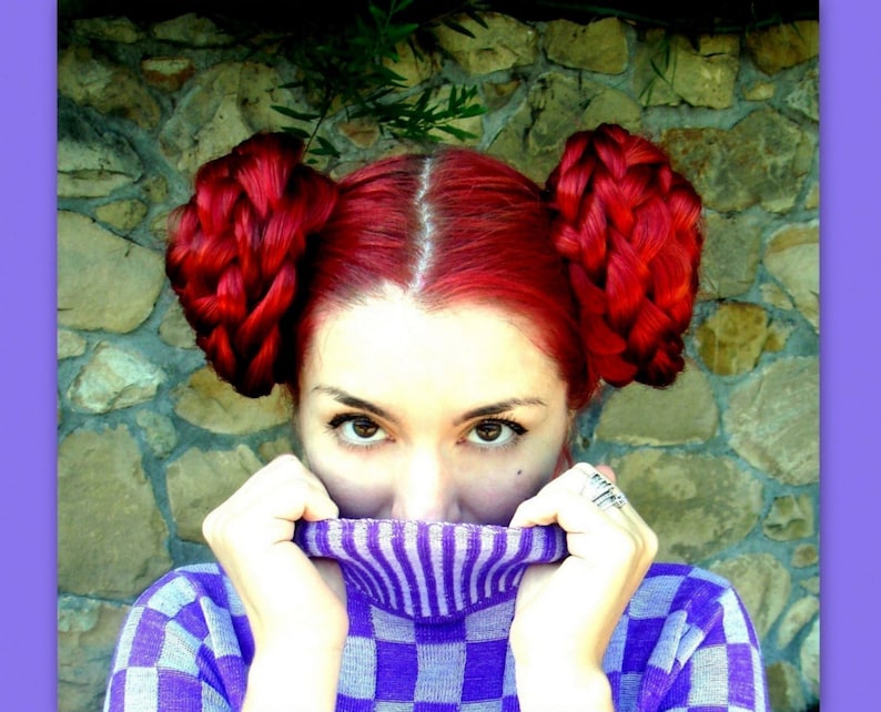 May include: A woman with bright red hair styled in two braided buns, wearing a purple and white checkered sweater with a purple turtleneck.