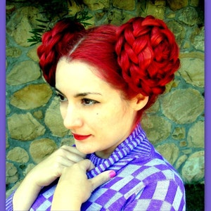 May include: A woman with bright red hair styled in two braided buns on either side of her head. She is wearing a purple and white checkered sweater.