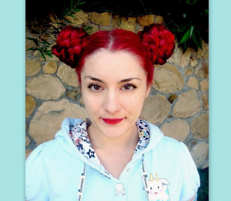 Puede incluir: Una persona con cabello rojo brillante peinado en dos mo&ntilde;os trenzados. Lleva una sudadera con capucha azul claro con un cuello estampado y un dibujo animado. La persona tiene l&aacute;piz labial rojo y est&aacute; mirando a la c&aacute;mara.