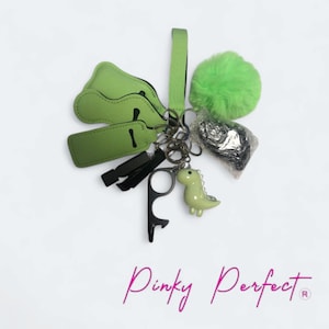 May include: A selection of keychains and accessories in green and black. Features a green dinosaur charm, a fluffy green pom-pom, and a bottle opener. The text "Pinky Perfect" is at the bottom.