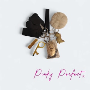 May include: A keyring set featuring a beige faux fur pom-pom, a small tan dinosaur charm, a gold-toned whistle, a gold-toned door opener tool, a gold-toned alarm device in a plastic bag, and three black fabric sleeves. The text Pinky Perfect is below.