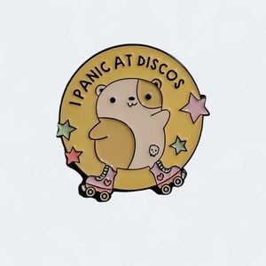 May include: Enamel pin with a cartoon hamster on roller skates. The hamster is tan and white, with a skull on its side. The pin is set against a yellow background with the text "I Panic at Discos" and star accents.