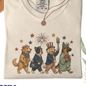 Puede incluir: Camiseta Comfort Colors de color marfil con un diseño de desfile de perros patrióticos. Los perros, incluyendo un golden retriever, un bulldog francés y un pastor alemán, visten atuendos festivos. La camiseta tiene el texto "Comfort Colors" y "XL-TG-EG".