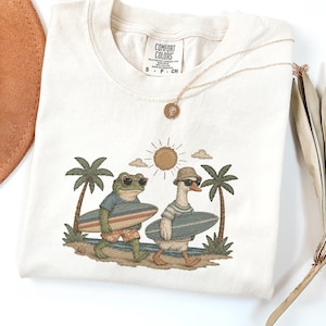 May include: Cream-colored Comfort Colors t-shirt featuring an embroidered design of a frog and a goose, both wearing sunglasses and carrying surfboards. The design includes palm trees, a sun, and clouds. A gold necklace and a brown hat are also visible.