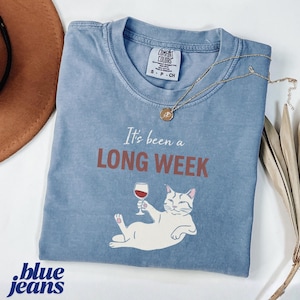 May include: A light blue Comfort Colors t-shirt with the phrase "It's been a LONG WEEK" and a graphic of a white cat holding a wine glass. A gold necklace and a brown hat are also visible.