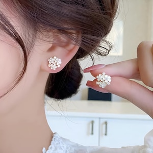 May include: Close-up of a pair of small, white flower-shaped stud earrings. One earring is in the ear, and the other is held up by a hand. The earrings have a cluster of small, delicate petals, creating a floral design.