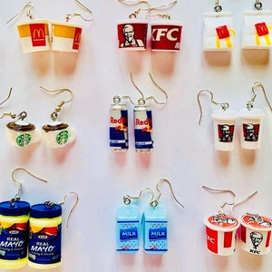 Könnte beinhalten: Eine Sammlung von ausgefallenen Ohrringen mit Miniatur-Lebensmitteln und -Getränken. Designs umfassen Burger, Pizzastücke, McDonald's- und KFC-Behälter, Starbucks-Tassen, Red Bull-Dosen, Milchkartons und Mayonnaise-Gläser.