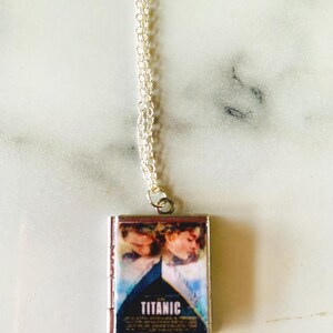 May include: A silver-toned necklace with a rectangular pendant featuring the movie poster for "Titanic." The pendant displays a blue and white image of the film's stars and the title "TITANIC" in white lettering. The chain is delicate and silver.