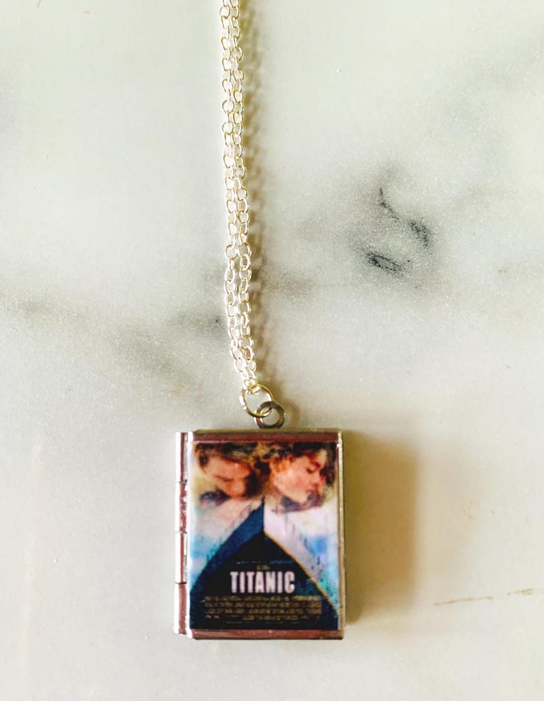 May include: A silver-toned necklace with a rectangular pendant featuring the movie poster for "Titanic." The pendant displays a blue and white image of the film's stars and the title "TITANIC" in white lettering. The chain is delicate and silver.