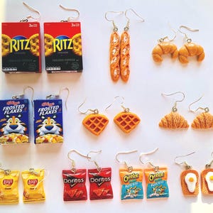 Peut inclure: Une collection de boucles d'oreilles fantaisie représentant des aliments miniatures. Les boucles d'oreilles comprennent des crackers Ritz, des baguettes, des croissants, des boîtes de céréales Frosted Flakes, des gaufres, des chips Lay's, des Doritos, des Cheetos et des œufs au plat sur du pain grillé.