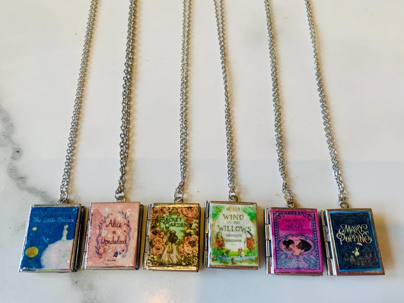 May include: Six silver-toned necklaces each feature a miniature book pendant. The book covers display titles like "The Little Prince," "Alice in Wonderland," and "Mary Poppins." The pendants are rectangular with hinged openings.