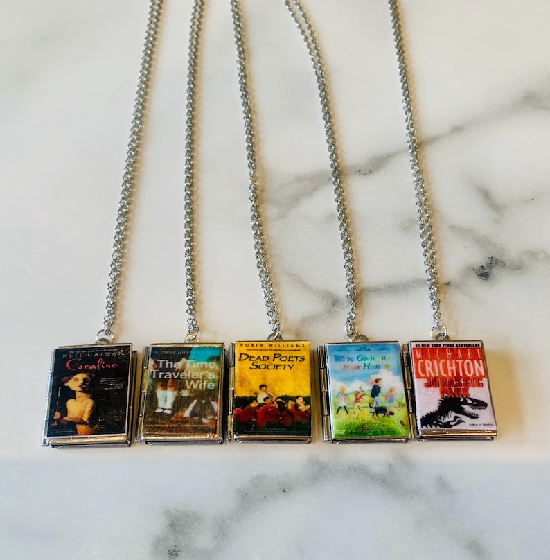 May include: Five silver chain necklaces, each with a miniature book pendant. The book covers feature titles like "Coraline" and "The Time Traveler's Wife." The pendants are rectangular with detailed book cover art.