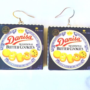 K&ouml;nnte beinhalten: Quadratische Ohrringe mit dem Design der Danisa Butterkeks-Box. Der Text "Danisa Traditional Butter Cookies" ist in Rot und Wei&szlig; auf dunklem Blau zu sehen. Die Ohrringe haben silberfarbene Haken.