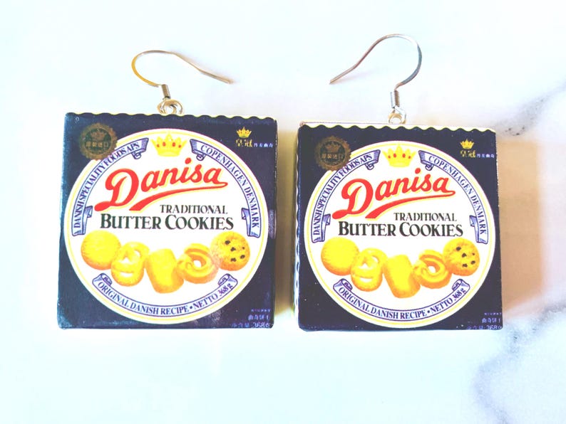 K&ouml;nnte beinhalten: Quadratische Ohrringe mit dem Design der Danisa Butterkeks-Box. Der Text "Danisa Traditional Butter Cookies" ist in Rot und Wei&szlig; auf dunklem Blau zu sehen. Die Ohrringe haben silberfarbene Haken.