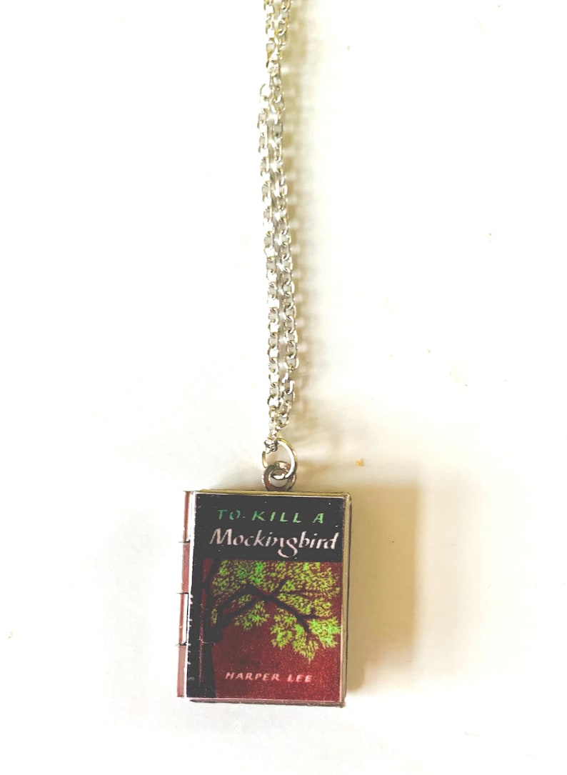 May include: A silver-colored chain necklace with a miniature book pendant. The book cover features the title "To Kill a Mockingbird" with a tree illustration and the author's name, Harper Lee, on a red background.